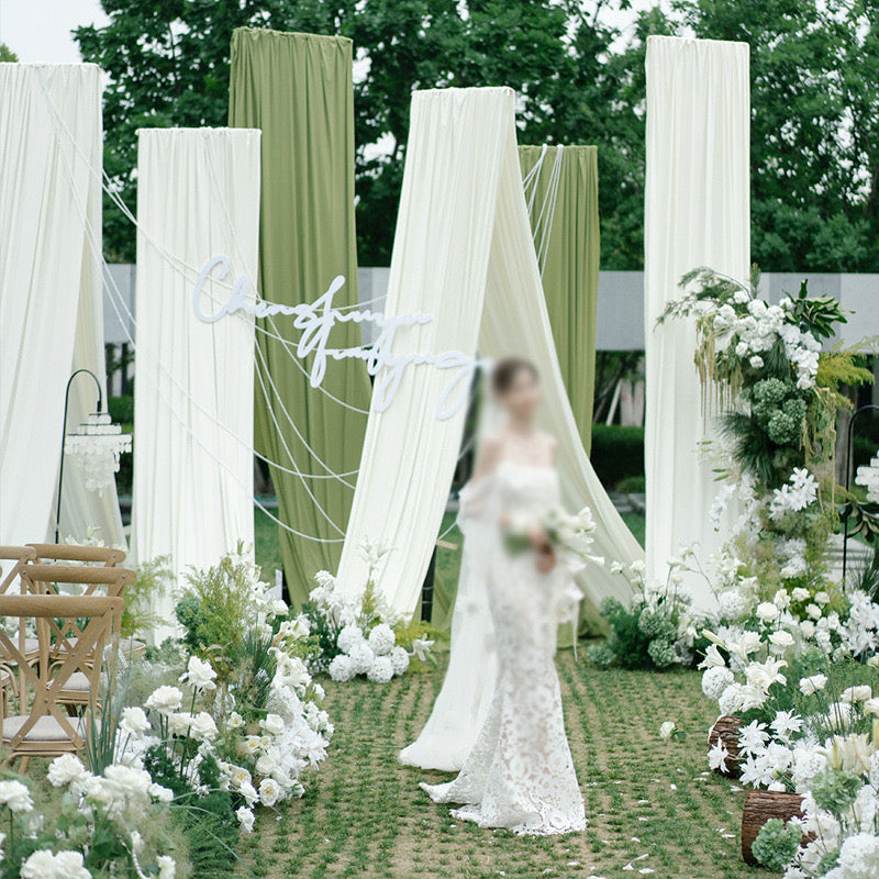 Elegant drapery arch stand with white fabric and floral decorations set up outdoors for a wedding ceremony.