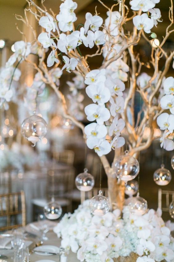 Elegant silk orchid centerpiece with gold branches and hanging orbs with tealight candles, perfect for weddings and events.