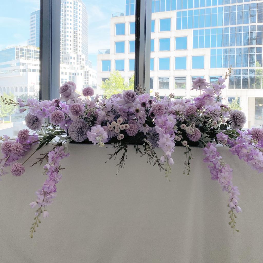Beautiful Rectangle Silk Floral Arrangement in shades of purple and white, displayed on a table against a cityscape backdrop.